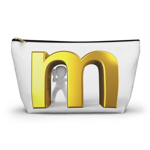 May include: White cosmetic bag with a gold zipper and a large gold letter "m" on the front. A small white figure is standing inside the letter. The bag is designed to hold makeup and other beauty products.