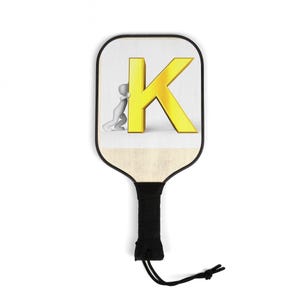Könnte beinhalten: Pickleball-Schläger mit schwarzem Griff und weißer Oberfläche. Auf der Oberfläche befindet sich ein goldener Buchstabe "K" mit einer kleinen Figur, die dagegen drückt. Eine schwarze Kordel mit Knoten ist am Griff befestigt.