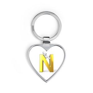 Op de afbeelding: Een zilveren hartvormige sleutelhanger met een gouden letter "N" erin. Een klein wit figuur klimt op de letter. De sleutelhanger heeft een zilveren ring aan de bovenkant om sleutels aan te bevestigen.