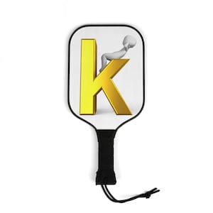 Op de afbeelding: Pickleball paddle met een witte voorkant met een gouden letter "K" en een kleine grijze figuur. De paddle heeft een zwart handvat met een zwarte polsband. De paddle is ontworpen voor pickleball.