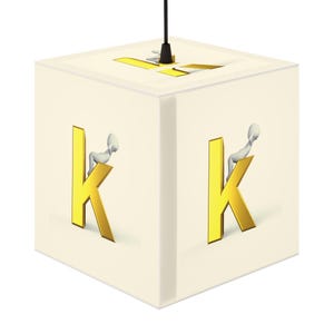 Op de afbeelding: Een vierkante, crèmekleurige hanglamp met een zwart snoer. De lamp heeft de letter "K" in goud op drie zijden, met een klein, wit figuur op elke "K". De bovenkant van de lamp heeft een gouden ontwerp.
