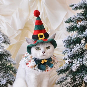 Puede incluir: Un gato blanco con un gorro de elfo verde y rojo con un collar de encaje blanco y un lazo verde. El gato está sentado frente a dos pequeños árboles de Navidad.