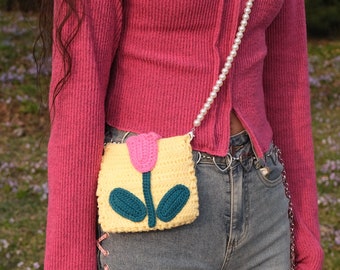 May include: A pink and yellow crocheted flower-shaped purse with a pearl chain strap. The purse is worn over a pink sweater and blue jeans.