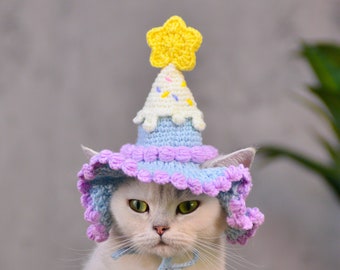 May include: A white cat wearing a blue and purple crocheted hat with a yellow star on top. The hat is shaped like a birthday cake.