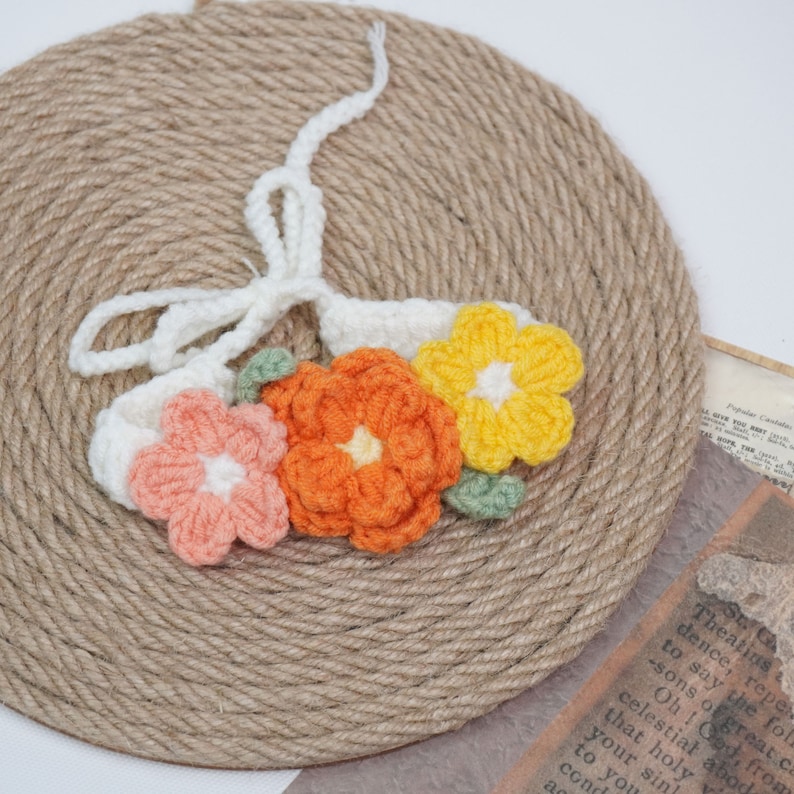 May include: A white crocheted headband with three crocheted flowers. The flowers are orange, yellow, and pink.