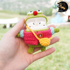 Pode incluir: Um sapo de croch&ecirc; verde vestindo um su&eacute;ter rosa e uma bolsa amarela, segurando um estojo branco para AirPod.