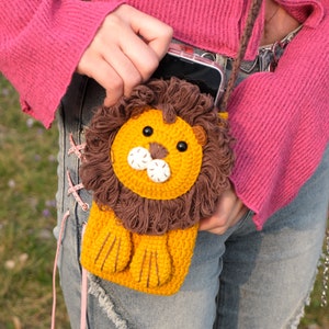 May include: A crocheted lion phone case with a brown mane and yellow face. The case is attached to a pink sweater and blue jeans.