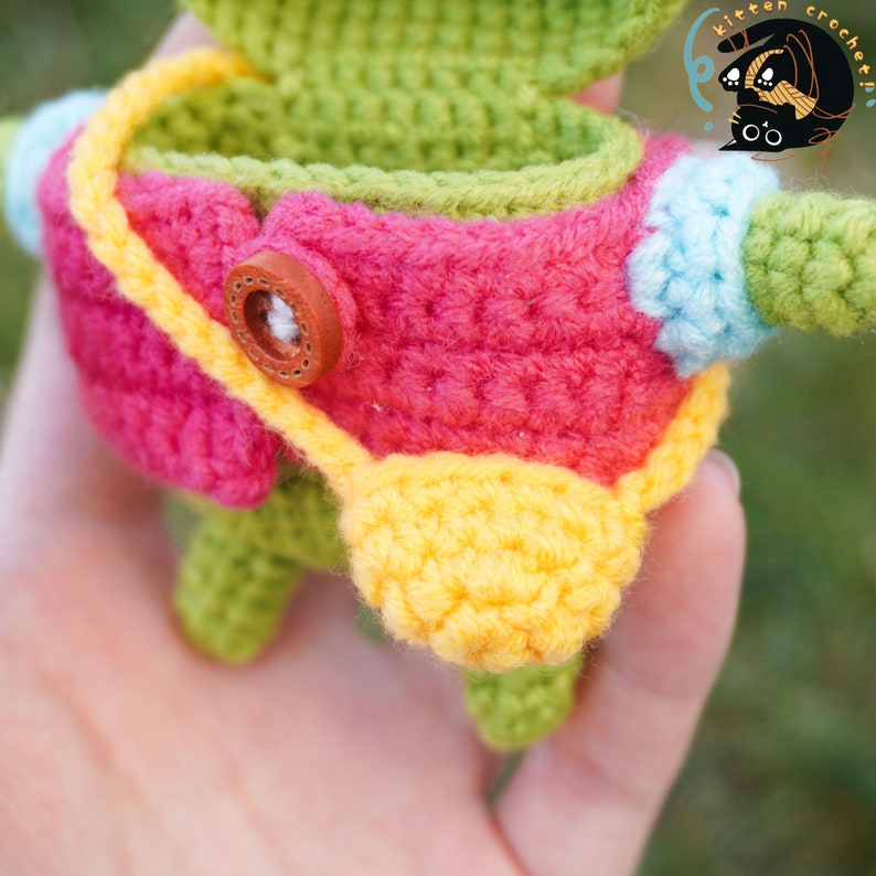 Pode incluir: Um brinquedo amigurumi de croch&ecirc; com um corpo verde, roupas rosa e amarelas e um bot&atilde;o marrom. O brinquedo est&aacute; sendo segurado em uma m&atilde;o.