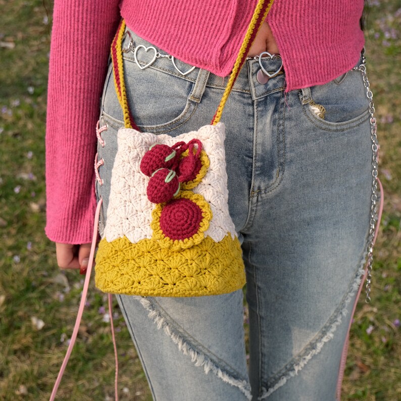 May include: A crocheted bag with a white and yellow design. The bag has a red flower and three red cherries on the front. The bag is being worn by a person wearing a pink sweater and blue jeans.