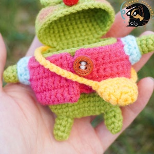 Pode incluir: Um sapo verde de croch&ecirc; com uma jaqueta rosa, uma al&ccedil;a amarela e um bot&atilde;o marrom. A boca do sapo est&aacute; aberta e o interior do sapo &eacute; vis&iacute;vel.