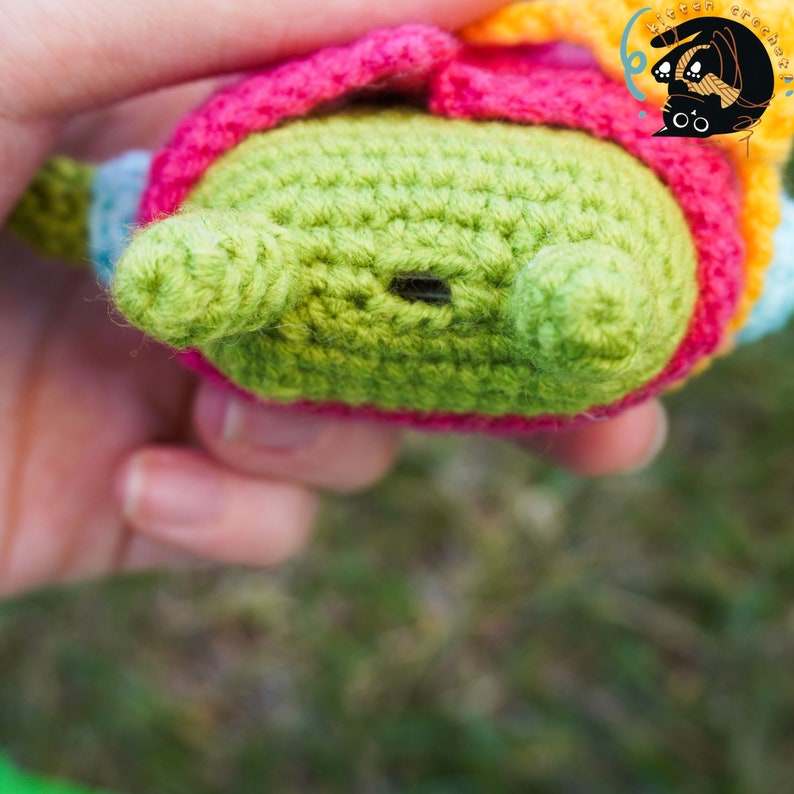 Pode incluir: Um brinquedo amigurumi de croch&ecirc; com um corpo verde, detalhes rosa e amarelos e um nariz de bot&atilde;o preto. O brinquedo est&aacute; sendo segurado por uma m&atilde;o.