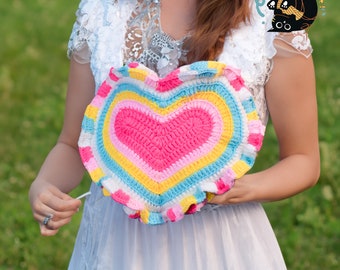 May include: A woman holds a crocheted heart-shaped pillow with a pink center and rainbow stripes. The pillow has a ruffled edge.