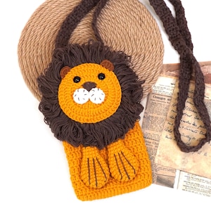 May include: A crocheted lion-shaped purse with a brown strap. The purse is orange and brown with a white nose and black eyes.