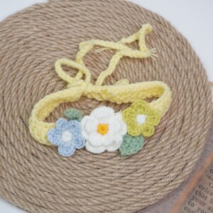 May include: A yellow crocheted headband with three crocheted flowers. The flowers are white, blue, and green.