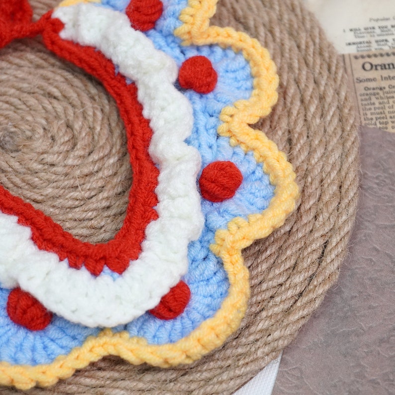 May include: A crocheted collar with a scalloped edge in blue, white, and yellow with red pom-poms.