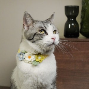 May include: A white and gray cat wearing a crocheted flower collar with yellow, green, and white flowers.
