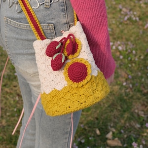 May include: A crocheted handbag with a pink and white base and a yellow top. The bag is decorated with crocheted cherries and a red flower.