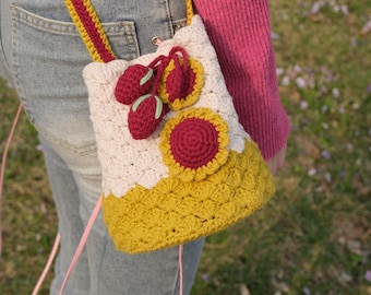 May include: A crocheted handbag with a pink and white base and a yellow top. The bag is decorated with crocheted cherries and a red flower.