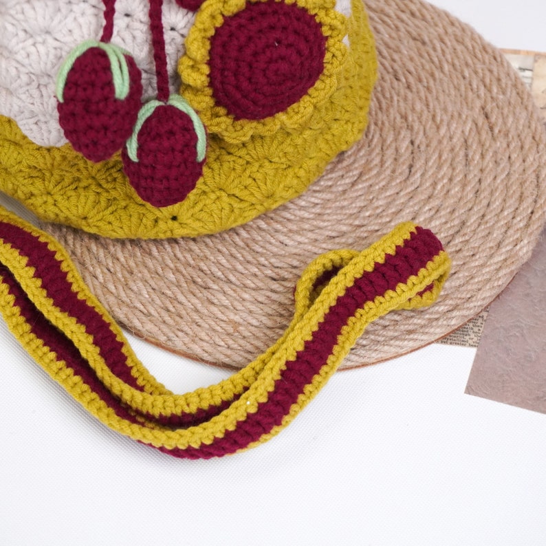 May include: A crocheted bag with a mustard yellow and burgundy striped strap. The bag is made of a beige, white, and burgundy yarn and features a burgundy flower with a yellow center.