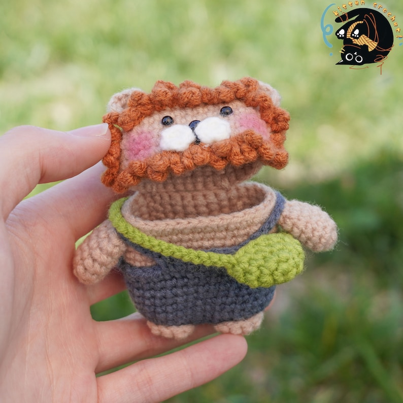 Pode incluir: Um brinquedo de le&atilde;o de croch&ecirc; com uma juba remov&iacute;vel, vestindo um macac&atilde;o e uma mochila verde. O brinquedo &eacute; marrom, laranja e verde.