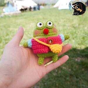 Pode incluir: Um sapo de croch&ecirc; verde vestindo um colete de croch&ecirc; rosa com uma bolsa de croch&ecirc; amarela. O sapo tem um nariz vermelho e olhos brancos.