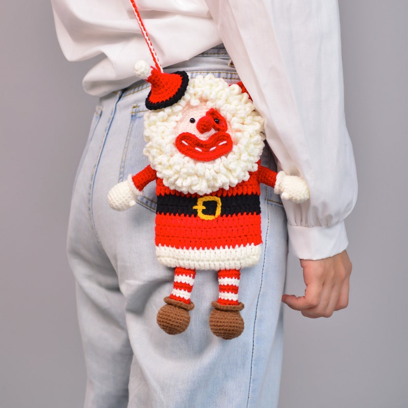 Pu&ograve; includere: Un ornamento di Babbo Natale all'uncinetto con una barba bianca, gambe a righe rosse e bianche e una cintura nera. L'ornamento &egrave; attaccato a una camicia bianca con jeans blu.
