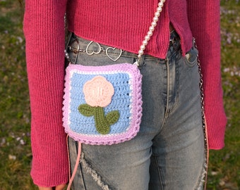 May include: A small crocheted bag with a pink flower design. The bag is light blue with a white border and a pink flower in the center. The bag is attached to a pearl chain.