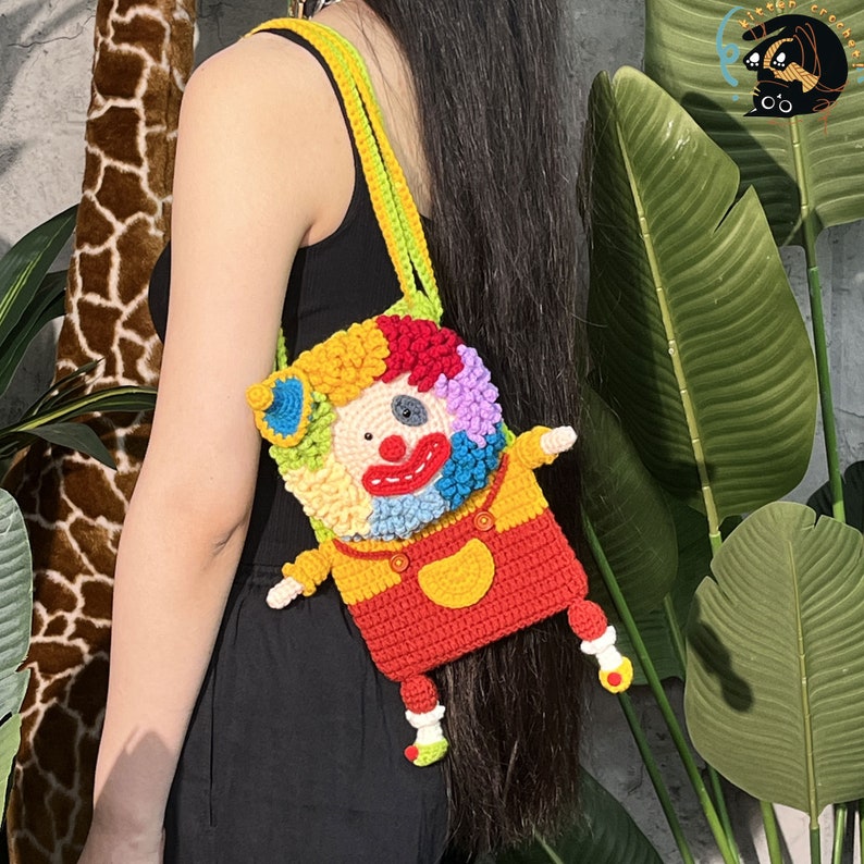 Pu&ograve; includere: Una borsa a mano da clown colorata all'uncinetto con un viso rosso, giallo e blu. La borsa ha una tracolla verde ed &egrave; indossata da una persona.
