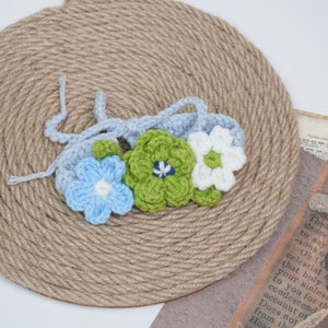 May include: A crocheted headband with three flowers in blue, green, and white. The headband is made of soft yarn and is perfect for newborn photography.