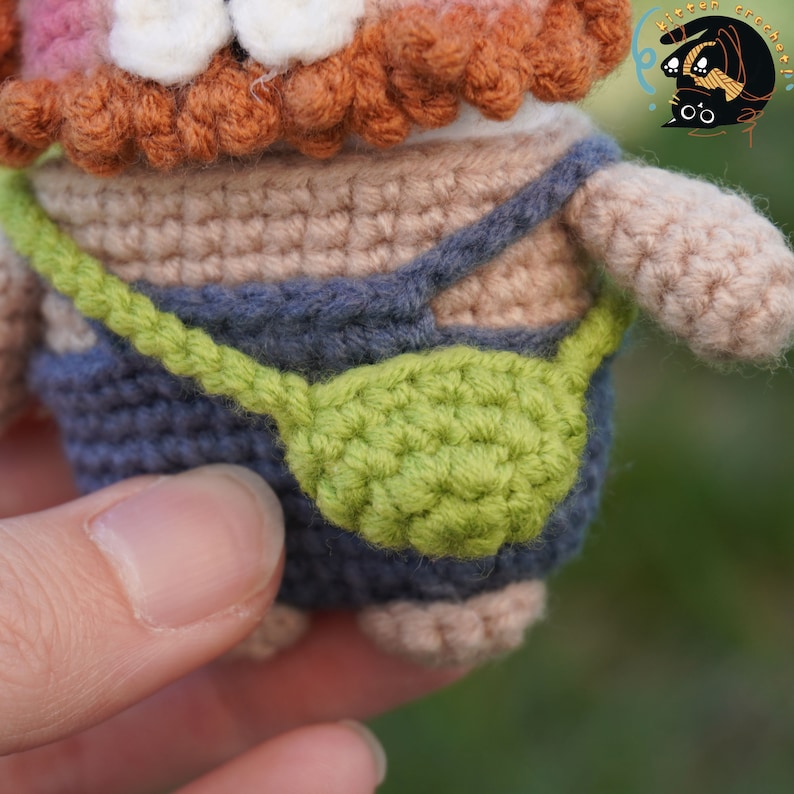 Pode incluir: Um brinquedo de amigurumi de croch&ecirc; com barba marrom, vestindo um macac&atilde;o azul com uma bolsa verde. O brinquedo est&aacute; sendo segurado por uma m&atilde;o.
