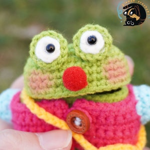 Pode incluir: Um sapo de croch&ecirc; verde vestindo uma jaqueta de croch&ecirc; rosa e amarela com um bot&atilde;o marrom. O sapo tem olhos grandes brancos com pupilas pretas e um nariz vermelho.