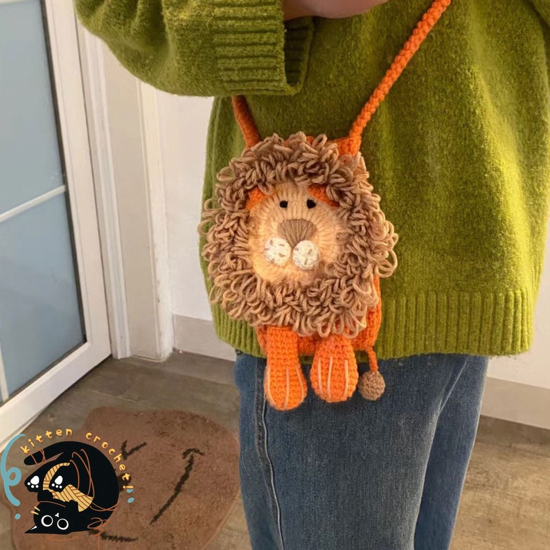 May include: A crocheted lion-shaped purse with orange and brown fur. The purse is worn as a crossbody bag by a person wearing a green sweater and blue jeans.
