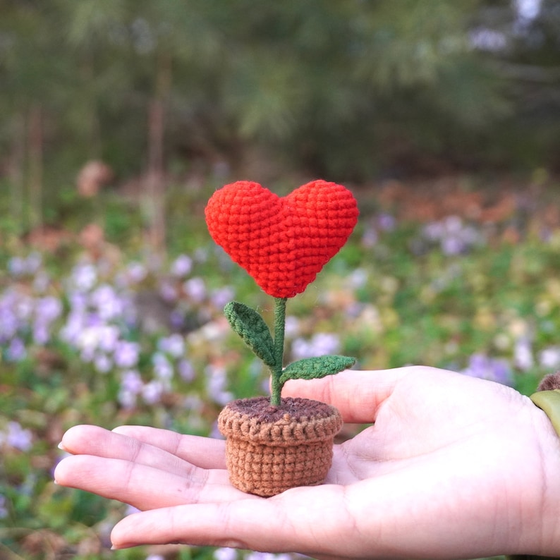 Op de afbeelding: Een gehaakte bloem in de vorm van een hart in een bruine pot, vastgehouden in een hand tegen een wazige achtergrond van groen gebladerte en paarse bloemen.