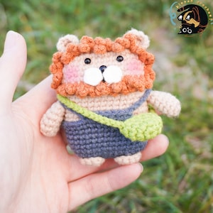 Pode incluir: Um brinquedo de le&atilde;o de croch&ecirc; vestindo um macac&atilde;o azul e uma bolsa verde. O le&atilde;o tem cabelo laranja, um focinho branco e olhos castanhos.