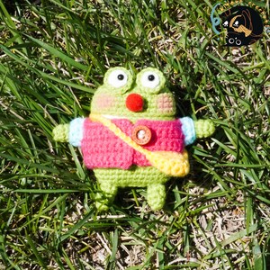 Pode incluir: Um sapo de croch&ecirc; verde vestindo uma jaqueta de croch&ecirc; rosa e amarela com um bot&atilde;o marrom. O sapo est&aacute; sentado em um leito de grama verde.