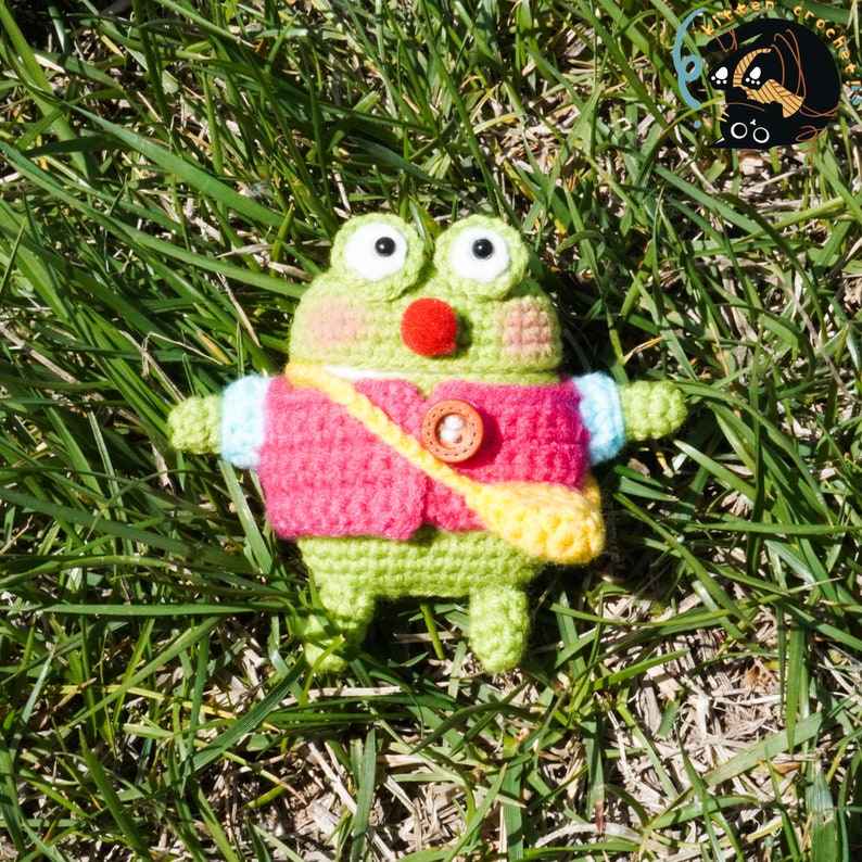 Pode incluir: Um sapo de croch&ecirc; verde vestindo uma jaqueta de croch&ecirc; rosa e amarela com um bot&atilde;o marrom. O sapo est&aacute; sentado em um leito de grama verde.