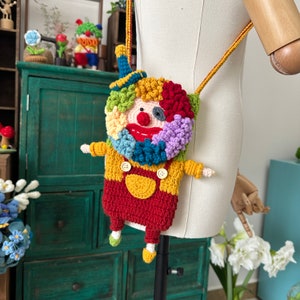 Pu&ograve; includere: Una borsa a mano da clown colorata all'uncinetto con un naso rosso, bottoni gialli e un cappello blu. La borsa &egrave; attaccata a una lunga tracolla.