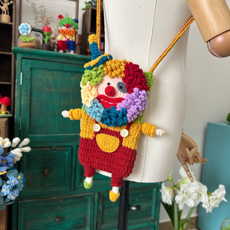 Pu&ograve; includere: Una borsa a mano da clown colorata all'uncinetto con un naso rosso, bottoni gialli e un cappello blu. La borsa &egrave; attaccata a una lunga tracolla.