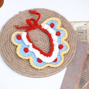 May include: A crocheted collar with a red, white, and blue color scheme. The collar has a scalloped edge and red polka dots. The collar is tied with a red ribbon.