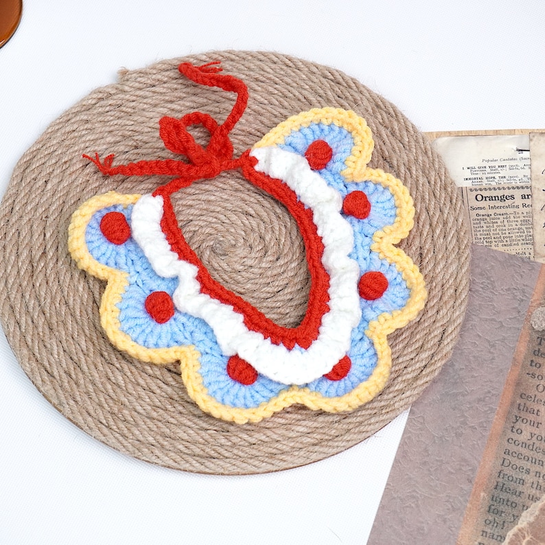 May include: A crocheted collar with a red, white, and blue color scheme. The collar has a scalloped edge and red polka dots. The collar is tied with a red ribbon.