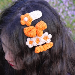 May include: A set of five crocheted hair clips in orange and white. The clips are shaped like flowers and bows.