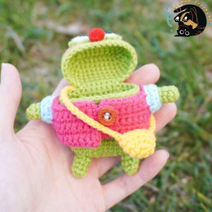 Pode incluir: Um brinquedo de sapo de croch&ecirc; com um nariz vermelho, um corpo verde, uma jaqueta rosa e uma al&ccedil;a amarela. A boca do sapo est&aacute; aberta, revelando o interior do brinquedo.