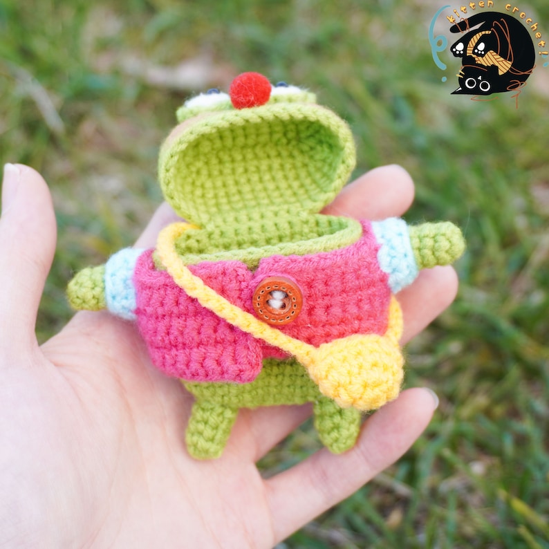 Pode incluir: Um brinquedo de sapo de croch&ecirc; com um nariz vermelho, um corpo verde, uma jaqueta rosa e uma al&ccedil;a amarela. A boca do sapo est&aacute; aberta, revelando o interior do brinquedo.