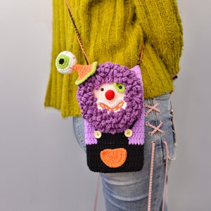 Pu&ograve; includere: Una borsa a tracolla in crochet viola e nera con un viso da clown e un solo occhio in cima. La borsa &egrave; indossata come una borsa a tracolla.