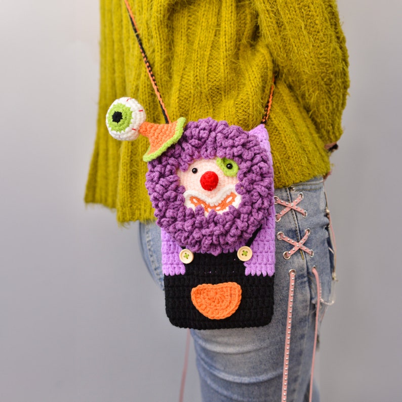 Pu&ograve; includere: Una borsa a tracolla in crochet viola e nera con un viso da clown e un solo occhio in cima. La borsa &egrave; indossata come una borsa a tracolla.