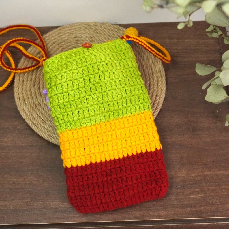 Pu&ograve; includere: Una custodia per telefono in crochet verde, giallo e rosso con un cinturino lungo. La custodia &egrave; realizzata in un filato morbido e testurizzato e ha un design semplice e moderno.