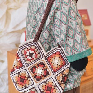 May include: A handmade crochet bag with a brown strap. The bag is composed of square granny squares in shades of brown, orange, and cream. The bag is carried over the shoulder. The background is blurred, but shows a person wearing a patterned shirt.