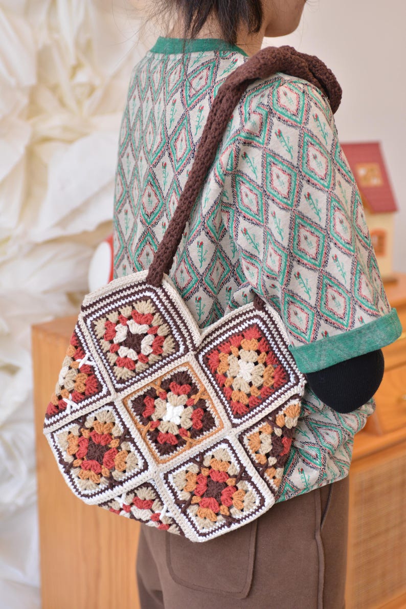 May include: A handmade crochet bag with a brown strap. The bag is composed of square granny squares in shades of brown, orange, and cream. The bag is carried over the shoulder. The background is blurred, but shows a person wearing a patterned shirt.