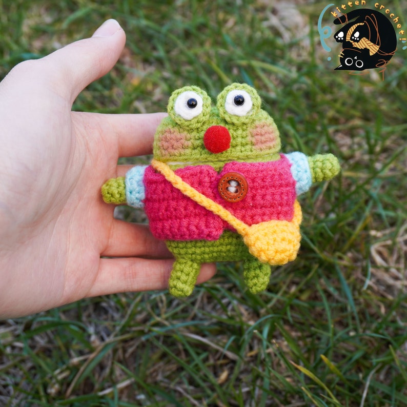 Pode incluir: Um sapo de croch&ecirc; verde vestindo um colete rosa e uma bolsa amarela. O sapo tem um nariz vermelho e olhos brancos.