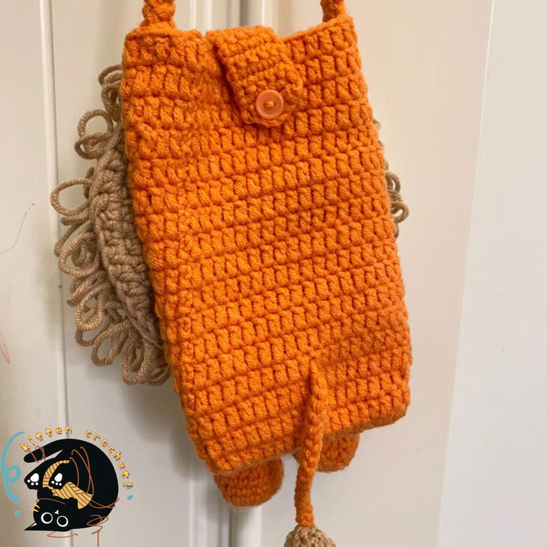 May include: Orange crocheted lion-shaped bag with a brown mane and a tail. The bag has a button closure and a strap.